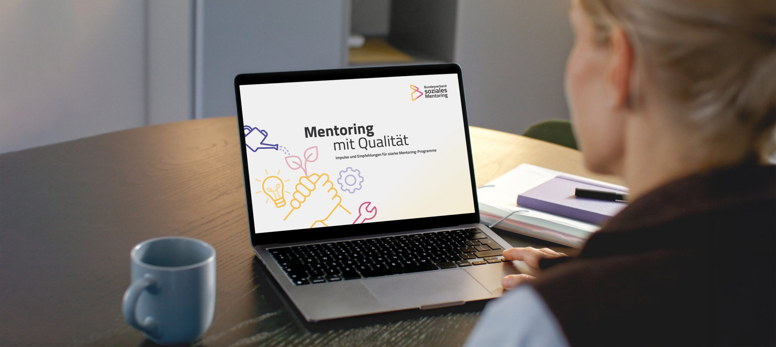 Mentoring mit Qualitaet Laptop-Mockup Person sitzt am Tisch vor einem Laptop, der das Cover der Publikation mit dem Titel "Mentoring mit Qualität" zeigt.